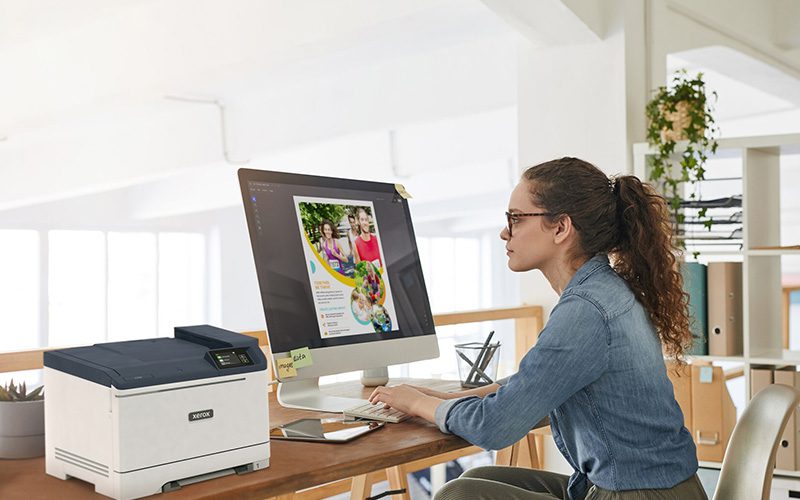 Une femme assise à un bureau, concentrée sur son travail, avec une imprimante Xerox® C320 couleur