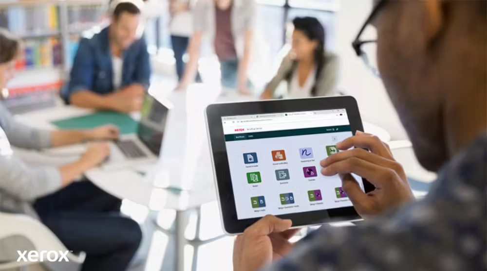 Plateforme Xerox® Workflow Central : accès mobile intuitif Une personne tient une tablette affichant l'interface Xerox Workflow Central avec diverses icônes d'application, tandis que d'autres personnes travaillent dans l'arrière-plan flou, mettant en évidence l'accès mobile aux solutions de flux de travail.