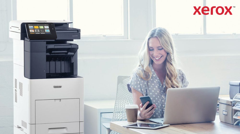 Xerox® ConnectKey® : intégration transparente des flux de travail Une personne regarde joyeusement son smartphone, assise à un bureau avec un ordinateur portable. Une imprimante multifonction Xerox VersaLink B605 est visible à côté du bureau, dans un espace de travail lumineux.