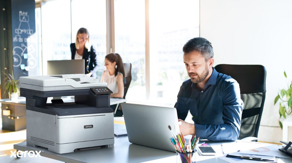 Xerox® Smart Start : conçu pour les espaces de travail modernes Une personne est assise à un bureau, les yeux rivés sur un ordinateur portable avec une imprimante multifonction Xerox C325 à côté. En arrière-plan, deux autres personnes travaillent également à leur bureau, dans un environnement lumineux.